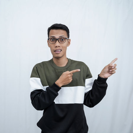 A young Asian man with glasses wearing a sweater smiles happily while pointing to the side of an empty space. Isolated on a white background.の写真素材