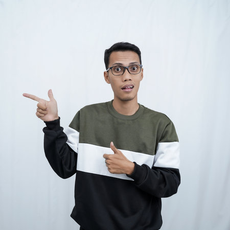 A young Asian man with glasses wearing a sweater smiles happily while pointing to the side of an empty space. Isolated on a white background.の写真素材