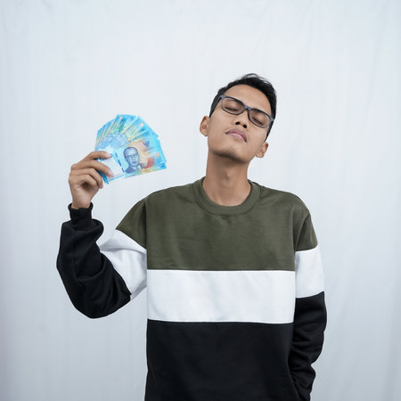 An asian young man with glasses wearing a sweater with a happy expression while carrying money, indonesian currency. Isolated on white background.の写真素材