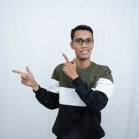 A young Asian man with glasses wearing a sweater smiles happily while pointing to the side of an empty space. Isolated on a white background.の写真素材