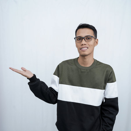 A young Asian man with glasses wearing a sweater smiles happily while pointing to the side of an empty space. Isolated on a white background.の写真素材