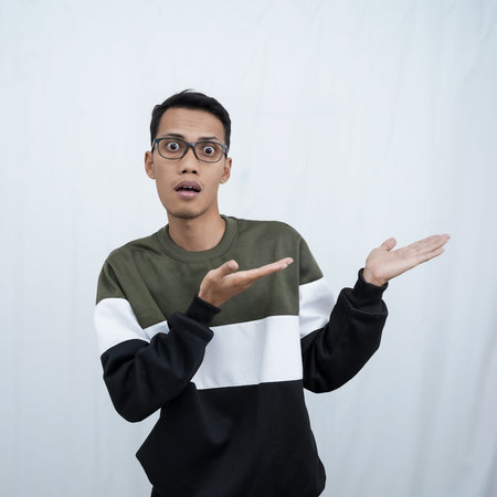 A young Asian man with glasses wearing a sweater with a shocked, surprised expression while pointing...