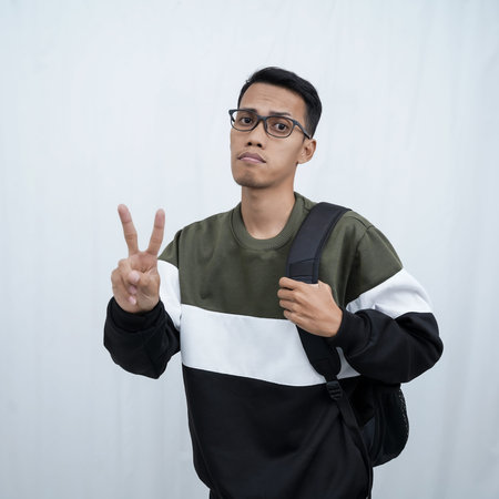 A young Asian man with glasses who is still in school wearing a sweater and a bag is posing standing...