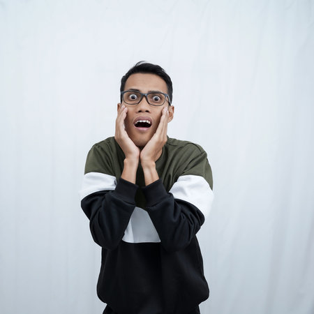A young asian man with glasses wearing a sweater with a shocked, surprised expression. Isolated on a...