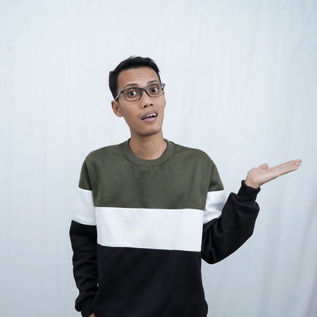 A young Asian man with glasses wearing a sweater smiles happily while pointing to the side of an...