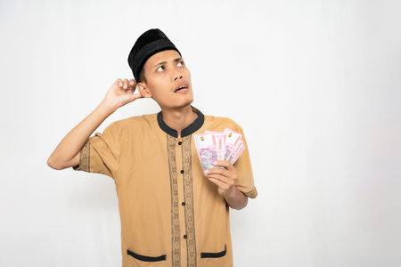 A young man in a brown shirt and black hat holding a stack of money and looking thoughtful