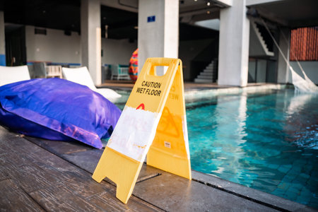 A yellow wet floor caution sign stands on a wooden poolside deck, warning of slipping hazards next...