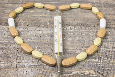 Glass thermometer, heart medications tablets the background of a wooden table. The view from the top