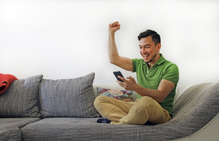 Man on couch excited by news on smartphoneの写真素材
