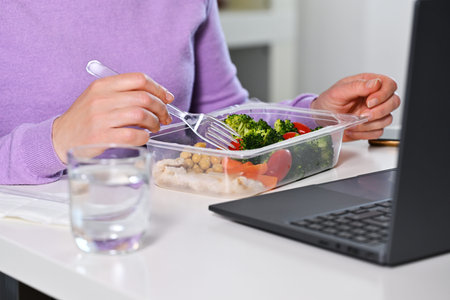 Woman having lunch from plastic box in front of laptop, closeup