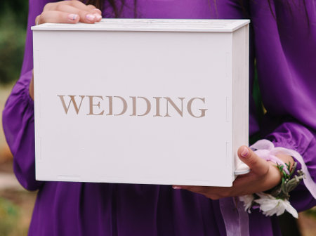 White box with sign Wedding. Bridesmaid in purple dress holds box for gifts. Outdoor lilac weddind ceremony. Invitatiaon box closeupの写真素材