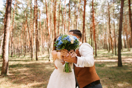 Autumn wedding at pine forest. Bride and groom kissing and holding wedding bouquet with blue flowers. Wedding in white, blue and brown colors.の写真素材