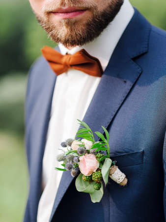 Bearded groom in a blue suit with a red bow tie and a boutonniere of dried flowers and tender roses. Groom's boutonniere closeup. Wedding inspiration. Fiance's fashionable look.の写真素材