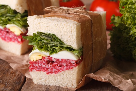 delicious homemade sandwich with salami, egg and greens wrapped in paper close-up on the tableの写真素材