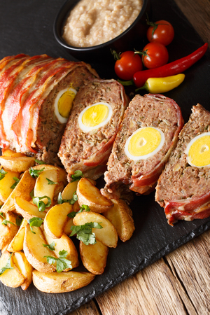 Baked meat loaf wrapped in bacon stuffed with egg and served with potato wedges and sauce close-up on a slate board on the table. verticalの写真素材