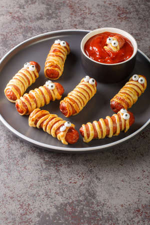 Scary mummy sausages in the dough with funny eyes on the table close-up. Funny decorations. Halloween food. Verticalの写真素材