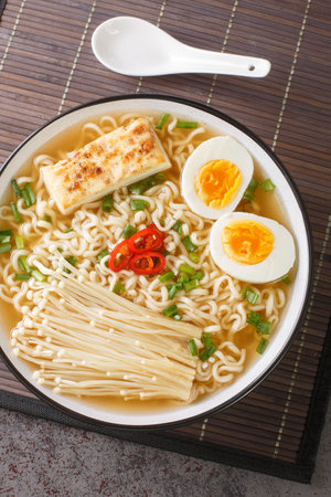Ramen soup with tofu cheese, enoki mushrooms, noodles, green onions and chili peppers close-up in a bowl on the table. Vertical top view from aboveの写真素材