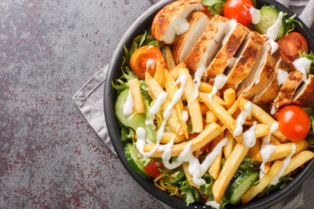 Pittsburgh steak salad consists of a lettuce salad with cheese, chicken breast, French fries, and ranch dressing on top closeup on the bowl on the table. Horizontal top view from aboveの写真素材