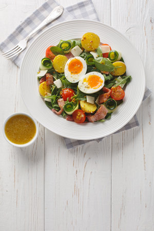 Fresh peasant salad with arugula, spinach, potatoes, bacon, tomatoes, cheese and soft-boiled eggs close-up on a plate on the table. Vertical top view from aboveの写真素材