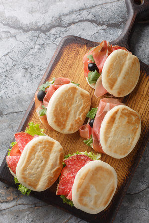 Popular street food in Emilia-Romagna, tigelle or crescentine sandwich with salami, ham and fresh vegetables close-up on wooden board on table. Vertical top view from aboveの写真素材