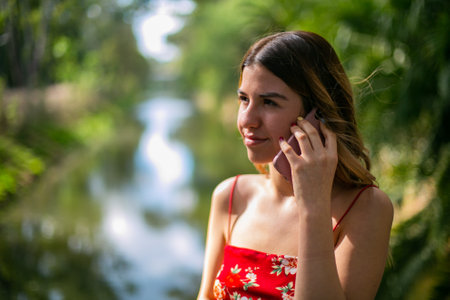 Beautiful young woman wears a red dress. She is talking on the phone in the parkの写真素材