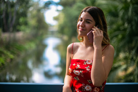Beautiful young woman wears a red dress. She is talking on the phone in the parkの写真素材