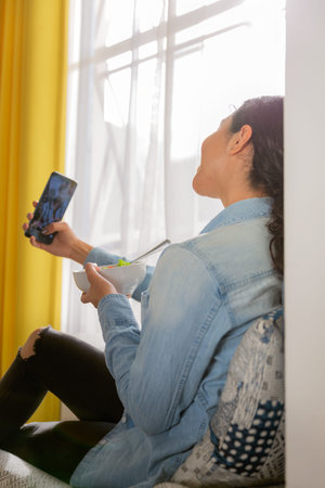 Smiling brunette young woman sitting on a chair and taking a selfie with her salad in her room