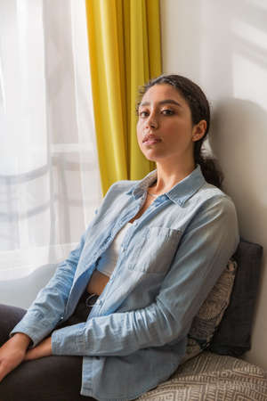 Portrait of a brunette woman sitting on a sofa in her room. Wear a denim shirt and black pants.の写真素材