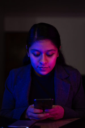 Night shot of a young woman working at home with her computer and tablet. She is using her phone.