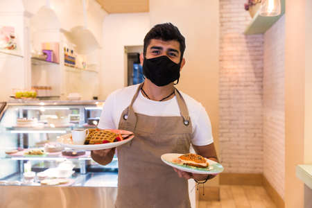 Portrait of a friendly waiter in a coffee shop. The background behind the waiter shows the interior of the cafeteria, he is wearing mask.の写真素材