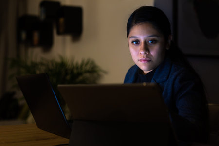Night shot of a young woman working at home with her computer and tablet
