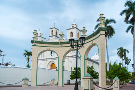 A white colonial church with twin bell towers stands next to a decorative arch in Pasaje La Concordia AhuachapÃ¡n El Salvador.の写真素材