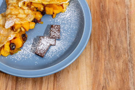 Mango crepes, with 3 pieces of chocolate, on a blue plate on a wooden table. Sugar Glass as...