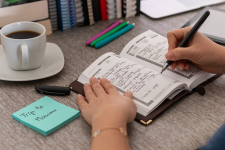 The woman writes something on a personal organizer. She is sitting at a desk. A laptop, a cup of...