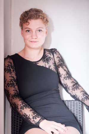 Pretty blonde ukrainian young woman with short hair sitting on a chair, dressed with black dress, against light background. Spainの写真素材