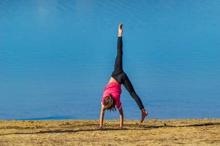 7 years old girl with pink shirt and sportswear doing a cartwheel at the beach during spring with blue water as background.の写真素材