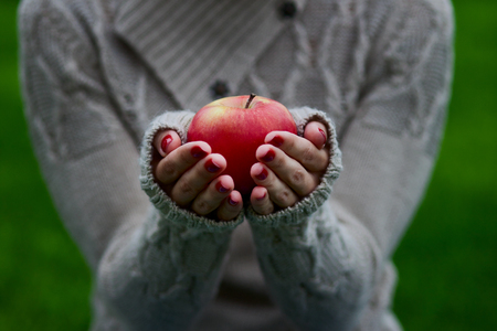 Closeup shot of a woman holding a red apple, with a green grass as background. A woman is wearing a gray sweaterの写真素材
