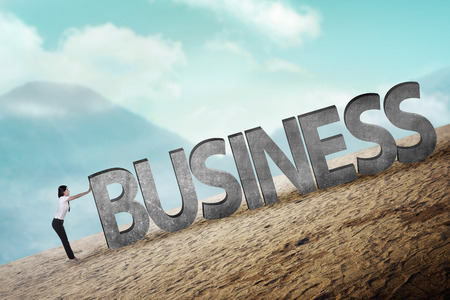 Asian business person pushing 3D business word uphillの写真素材