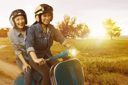 Two pretty asian woman having fun riding the scooter when sunset on the fieldの写真素材