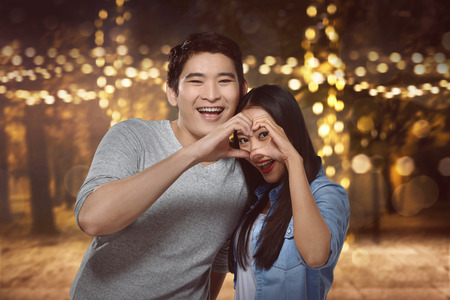 Asian couple standing close to each other and looking through a heart shape made with their handsの写真素材