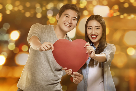 Happy asian couple holding handmade paper heart against blurred light backgroundの写真素材