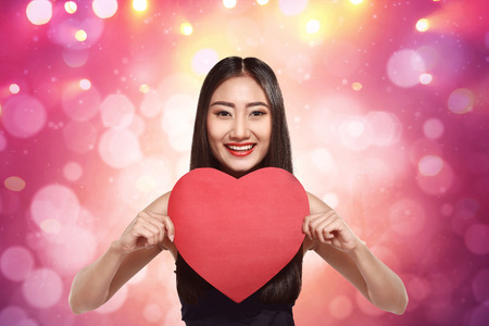 Smiling asian girl holding red heart shape against pink backgroundの写真素材