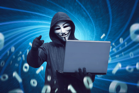 Hacker man with anonymous mask holding laptop against binary code in backgroundのeditorial素材