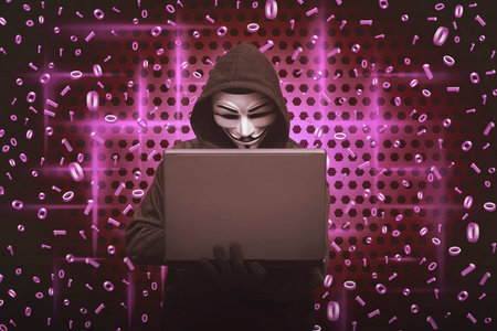 Hooded man wearing mask typing on laptop with binary code in background