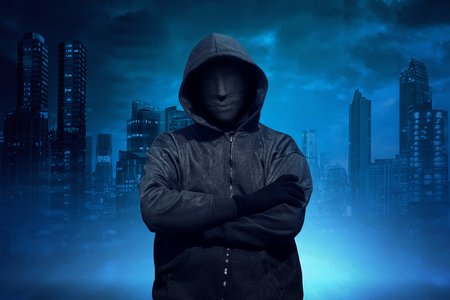 Hooded man with anonymous mask standing against cityscape backgroundの写真素材