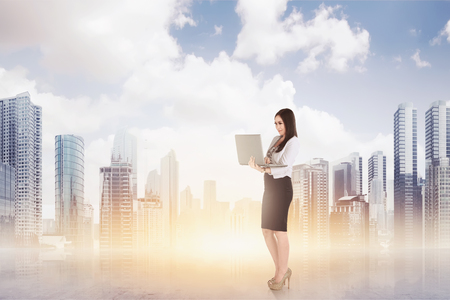 Attractive asian business woman using a laptop while standing against cityscape backgroundの写真素材