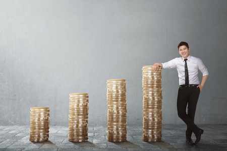 Confident asian businessman standing beside coin stack against grey wall backgroundの写真素材