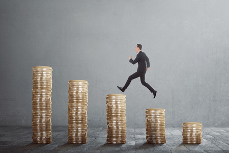 Happy asian businessman jump on between coin stacks against grey backgroundの写真素材