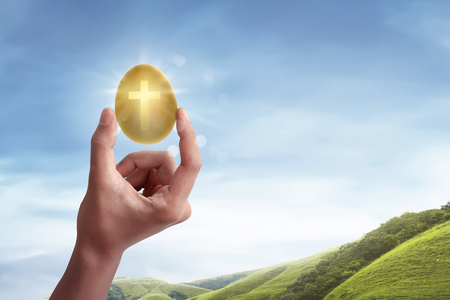 Hand holding the light of cross shape from the golden egg on the meadowの写真素材