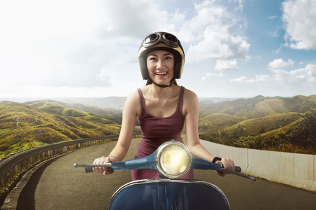 Happy asian woman riding blue scooter with helmet through mountain roadの写真素材